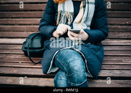 Close-up Junge unkenntlich Frau mit warmen Mantel sitzen auf einer Parkbank mit Smart Phone auf kalten Wintertag. Stockfoto