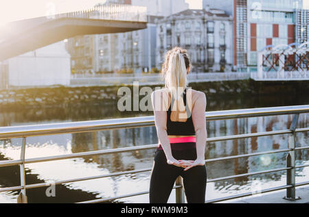 Rückansicht der jungen Dame in Sportswear stretching Hände am Kai in der Nähe von Wasser in der Stadt an einem sonnigen Tag Stockfoto