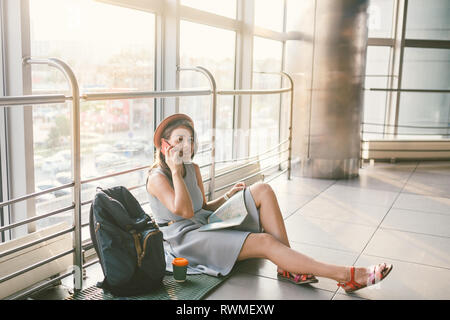 Thema Tourismus und Reisen von jungen Studenten. Schöne junge kaukasische Mädchen im Kleid und Hut sitzt auf dem Boden touristische Wolldecke im Terminal Flughafen Termin Stockfoto
