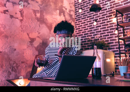 Junger Mann mit einem Glas Wein in der Hand in der Nähe des Tisches sitzen und einen Film auf dem Computer. Stockfoto