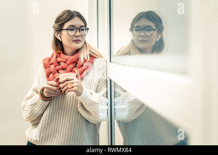 Friedliche langhaarigen Mädchen in die drahtlosen Kopfhörer suchen außerhalb des Fensters Stockfoto