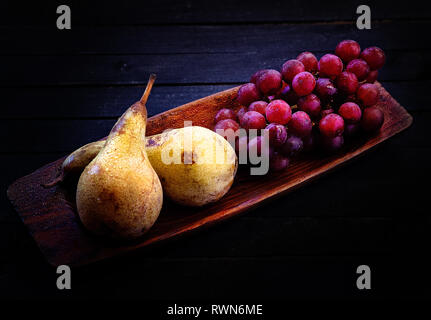Rote Trauben und Birnen. Holz- Sonnte sich auf einem Schwarzen Tisch. Reife Früchte. Wenig Licht Stockfoto