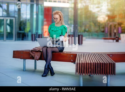 Junge Geschäftsfrau mit einem Laptop und trinken Kaffee auf einer Bank außerhalb des Bürogebäudes. Moderne Bürogebäude Stockfoto