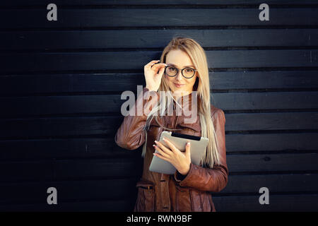 Blond Geschäftsfrau mit Tablette gegen schwarze Holz- Hintergrund. Tragen braune Lederjacke und Optische Brillen Stockfoto