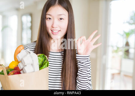 Schöne asiatische Frau mit papiertüte von frischen Lebensmitteln zu tun ok Zeichen mit den Fingern, ausgezeichnete Symbol Stockfoto