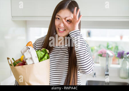 Schöne asiatische Frau mit papiertüte von frischen Lebensmitteln mit glücklichen Gesicht lächelnd tun ok Zeichen mit der Hand auf Auge schauen durch die Finger Stockfoto