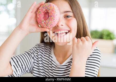 Schöne junge Mädchen Junge Süßigkeiten rosa Donut zeigen und zeigt mit dem Daumen bis zu der Seite mit glücklichen Gesicht lächelnd Stockfoto