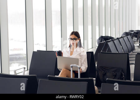 Informelle Besucherin Arbeiten am Laptop im Flughafen Halle. Frau seine Reise am Flughafen Terminal wartet, sitzt auf einem Stuhl und tippen auf dem Laptop. Stockfoto