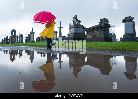 Frau in Gelb und Rot Regenschirm in Pfütze wandern in Nekropole Friedhof in Glasgow, Schottland, Großbritannien wider Stockfoto
