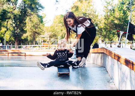 Junge Mutter lehrt, ihren kleinen Jungen ein Skateboard zu fahren Stockfoto