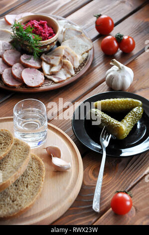 Ein Glas Wodka mit Essiggurken, Roggenbrot, frische Tomaten, Knoblauch, Speck, Wurst und geriebenem Meerrettich auf dem hölzernen Tisch, selektiver Fokus Stockfoto