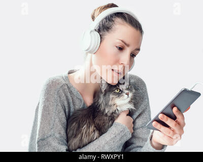 Stilvolle, junge Frau mit Kopfhörer, Handy und liebevolle, sanfte Kätzchen auf einem weißen Hintergrund, isoliert. Pet Care Concept Stockfoto