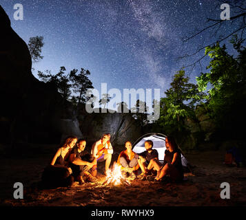 Gerne Freunde Wanderer mit einem Rest zusammen um Lagerfeuer neben Camp und beleuchtete touristische Zelt in der Nacht. Auf Hintergrund schöne Nacht sternenklaren Himmel voller Sterne und die Milchstraße, die Felsen. Stockfoto