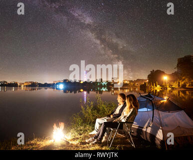 Nacht Camping am See in der Nähe von Lagerfeuer. Mann und Frau in eine Pause in der Nähe von Zelt. Paar sitzen auf Stühlen, genießen Sie einen wunderschönen Blick auf den nächtlichen Himmel voller Sterne und die Milchstraße, die Lichter der Stadt im Hintergrund. Stockfoto