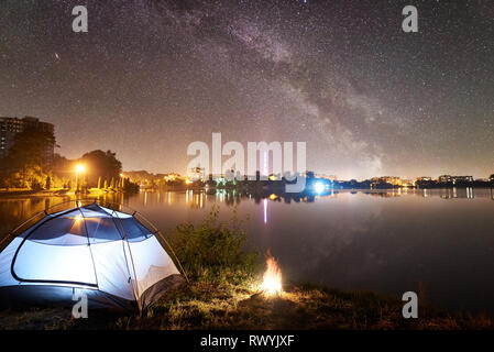 Nacht Camping. Beleuchtete touristische Zelt und Lagerfeuer auf dem Lake Shore unter schönen Abend Himmel voller Sterne und die Milchstraße, ruhige Wasseroberfläche, leuchtende Stadt auf Hintergrund. Outdoor Lifestyle Konzept Stockfoto