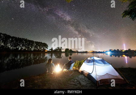 Nacht Camping auf See. Zurück Blick auf junge Familie - Mann und Frau sitzen auf Stühlen in der Nähe von Lagerfeuer und Zelt, Genießen am Abend Himmel voller Sterne, ruhige Wasserfläche, die Lichter der Stadt im Hintergrund. Stockfoto