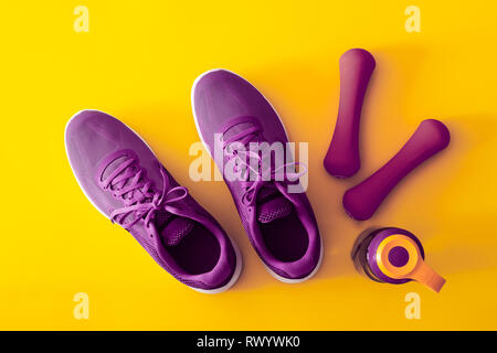 Blick von oben auf die lila und violett Sport Schuhe, Hanteln und eine Flasche Wasser auf gelben Hintergrund. Fitness und gesunde Lebensweise Hintergrund Konzept. Stockfoto