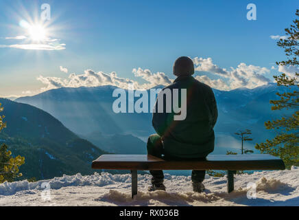 Mann allein sitzt auf einer Holzbank, mit Blick auf die schönen Berge. Rainbow Effekt auf schwarzen Mantel des Mannes. Stockfoto