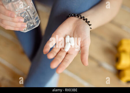 Sport Mädchen mit einem blauen Hemd und blau Leggings sitzt auf dem Boden und hält in ihren Händen sport Nahrungsergänzungsmittel in Kapseln für Bodybuilding Stockfoto