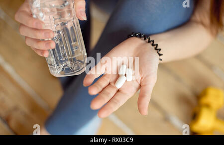 Sport Mädchen mit einem blauen Hemd und blau Leggings sitzt auf dem Boden und hält in ihren Händen sport Nahrungsergänzungsmittel in Kapseln für Bodybuilding Stockfoto