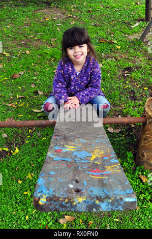 Wenig glücklich Mädchen mit einem Lächeln Spaß auf einem alten Holz abgeplatzte Farbe Wippe in einem sehr grünen Spielplatz im Herbst mit Blätter auf dem Boden Stockfoto