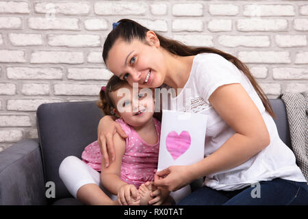 Lächelnde Mutter umarmte ihre kleine Tochter Holding Liebe Karte in der Hand zu Hause Stockfoto