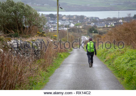 Ein Reisender geht die Straße in Richtung Dingle Stadt an einem kalten Tag im März sporting ein helles Grün Rucksack. Stockfoto