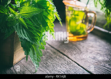 Bündel von brennnessel Pflanzen in einer Holzkiste und Glas von gesunden Brennnesseltee nicht im Fokus auf alten Holztisch im retro Haus im Dorf. Stockfoto