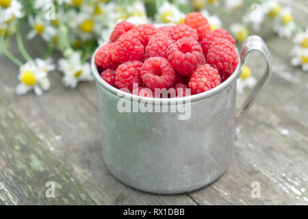 Vintage rustikalen Becher voller Himbeere Beeren auf Holztisch und Bündel von Daisy Blumen auf Hintergrund. Stockfoto