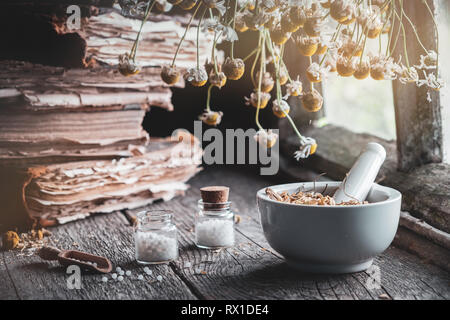 Mörtel von getrockneten Kräutern, Flaschen von homöopathischen Globuli, alte Bücher und Bündel trockenen Kamille Anlage. Homöopathische Medizin. Stockfoto