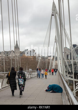 Touristen, ein Bettler auf einer der Goldenen Jubiläum Brücken über die Themse, London, UK, zu Charing Cross auf einem kalten, stürmischen Tag im März. Stockfoto