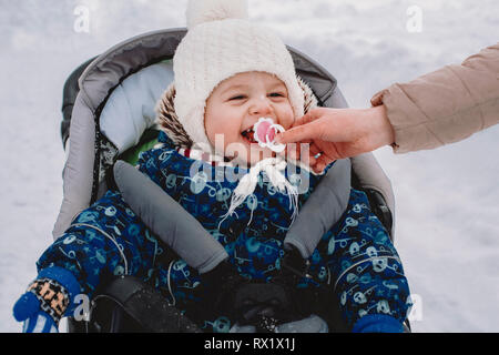 7/8 Hand der Mutter des Sohnes, Schnuller in Mund sitzen auf Baby Stroller im Park im Winter Stockfoto