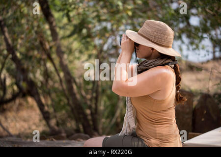 Seitenansicht der Frau durch ein Fernglas, während gegen Bäume im Wald sitzen Stockfoto