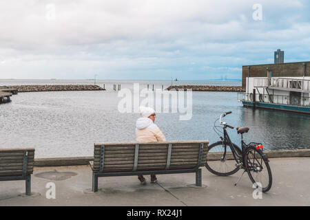 Eine junge Kaukasier Frau sitzt mit dem Rücken auf einer Holzbank mit Blick auf die Ostsee an der Küste in Kopenhagen Dänemark im Winter in trüben weat Stockfoto