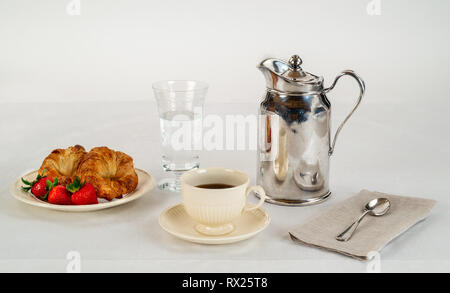 Cremefarbene Platte und Kaffee Tasse mit zwei Croissants und Drei frische Erdbeeren auf einem weißen Leinen Tischdecke und ein Silver Hotel Zimmer Service Karaffe Stockfoto