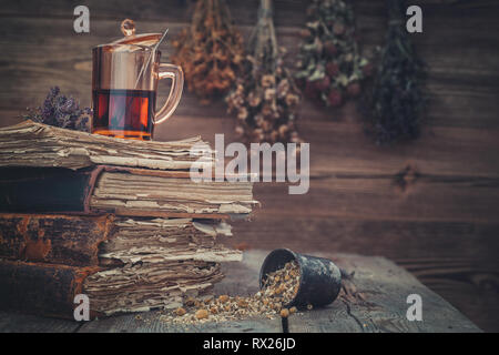 Cup der gesunden Tee oder Tinktur, Mörtel von gesunden daisy Kräuter und Stapel alter Bücher. Hängende Trauben von Heilpflanzen für den Hintergrund. Stockfoto