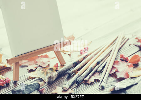 Leinwand auf Staffelei, malen Rohre, Bürsten für Malerei und Herbst Ahorn Blätter auf alten hölzernen Hintergrund. Retro Farben Foto. Stockfoto