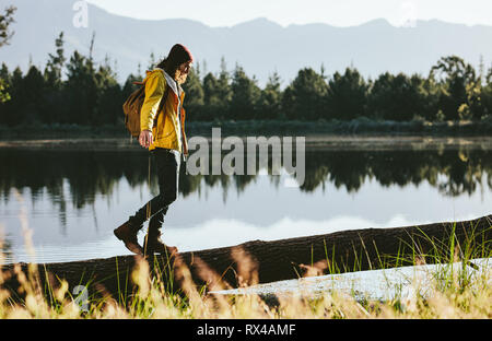 Touristische Mann zu Fuß auf einem gefallenen Baumstamm neben einem See. Mann mit Jacke und Rucksack wandern an einem See. Stockfoto