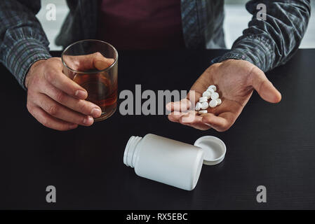 Zugeschnittenes Bild der männlichen Hände halten eine Pille jar und ein Glas Whiskey über schwarzen Tisch. Der Mensch leiden Kater in Alkoholismus und Alkoholmissbrauch. Ein Bott Stockfoto