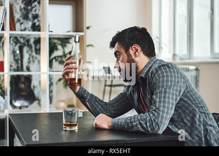 Trinken Mühe. Deprimiert bärtigen Mann sitzt am Tisch mit einer Flasche Whiskey in der Hand. Er schaut auf die Flasche während über Probleme denken nicht Stockfoto