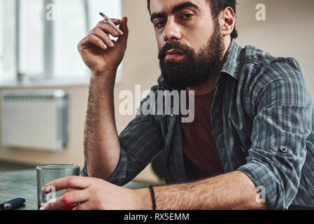 Zugeschnittenes Bild der Dunkelhaarige, traurig und verschwendet alkoholische Mann am Tisch sitzen, in der Küche, Rauchen und Trinken Whiskey, Glas und Zigarre Stockfoto
