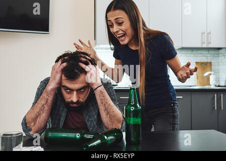 Betrunkener Mann. Dunkelhaarige süchtig Frau Gefühl der Verzweiflung und schrie ihren betrunkenen Mann in der Küche sitzt. Leere Flaschen auf dem Tisch Stockfoto