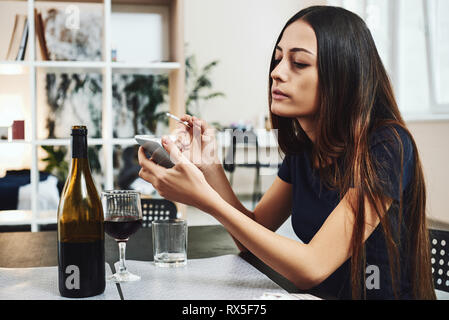 Porträt der jungen Frau, die in Verzweiflung, rauchen Zigarette während am Tisch sitzen, trinken Alkohol zu Hause. Sie wartet auf Anruf. Weibliche alcoholi Stockfoto