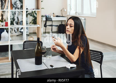 Porträt der jungen Frau, die in Verzweiflung, rauchen Zigarette während am Tisch sitzen, trinken Alkohol zu Hause. Sie wartet auf Anruf. Weibliche alcoholi Stockfoto