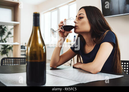 Deprimiert, geschiedene Frau sitzt allein in der Küche zu Hause und trinkt ein Glas Rotwein, da es Probleme bei der Arbeit und Mühen in Beziehungen. Stockfoto