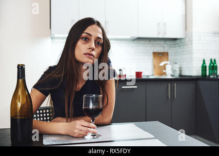 Dunkelhaarige, traurig und verschwendet alkoholische Frau zu Hause zu sitzen, in der Küche, trinken Rotwein, holding Glas, völlig betrunken, niedergedrückt, lo Stockfoto