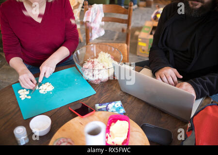 Blick hinunter auf eine sitzende Frau hacken Knoblauch und sitzenden Mann, der auf einem Laptop an einem Holztisch arbeitet Stockfoto