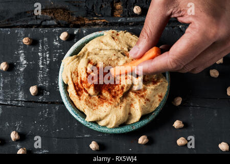 Hohe Betrachtungswinkel eines Mannes Eintauchen ein Streifen von Karotte in einem hausgemachten Hummus gewürzt mit Paprika in einer grünen Keramik Teller serviert, auf einem dunklen Grau rustikal w Stockfoto