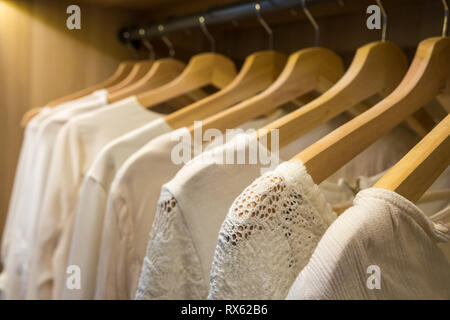Weiße Blusen und Pullover auf Kleiderbügel im Schrank mit Beleuchtung. Close-up. Stockfoto