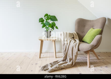 Beige Sessel mit einem grünen Kissen, Decke und einem Holztisch mit einer Topfpflanze, Obstsalat Baum (Monstera deliciosa). Leere weiße Wand in einfachen Stockfoto